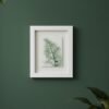 The Dill – Original Ceramic Wall Art, inspired by Irish design, features a delicate green leaf and is displayed on a dark green wall with leafy plants partially visible in the corners.