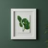 The Dill – Original Ceramic Wall Art, inspired by Irish design and featuring green leaves, is displayed in a white frame on a dark green wall with part of a leafy plant visible in the lower right corner.
