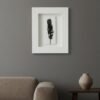 A minimalist living room with a beige sofa, a dark wooden table holding a simple beige vase, and Dill – Original Ceramic Wall Art featuring a black feather on the gray wall.