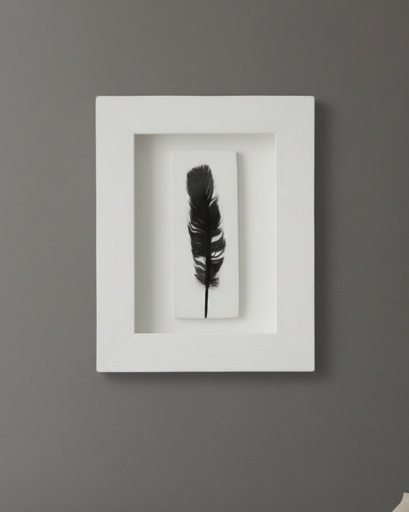 The Dill – Original Ceramic Wall Art features a single black feather centered on white, set in a white rectangular frame and displayed on a gray wall—a minimalist piece that captures the elegance of modern Irish design.