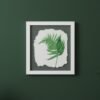 The Native Irish Fern Leaf – Original Ceramic Wall Art features a detailed ceramic fern leaf on textured white, elegantly framed in white against a dark green wall with hints of green leaves at the corners, enhancing its natural beauty.