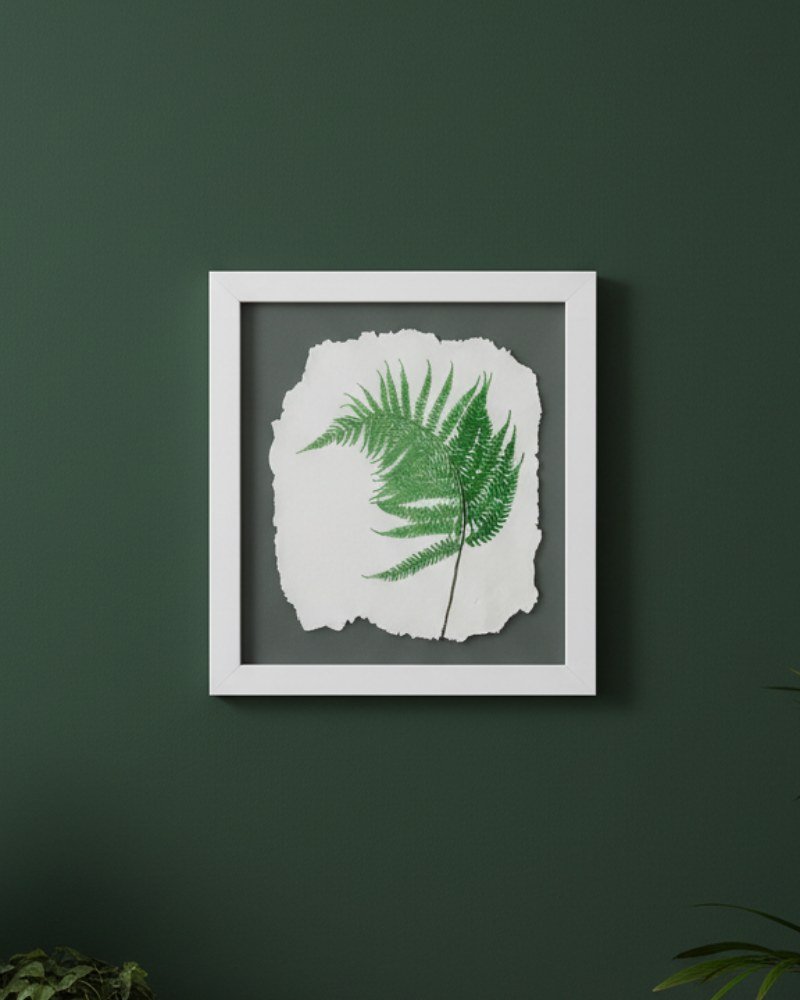 The Native Irish Fern Leaf – Original Ceramic Wall Art features a detailed ceramic fern leaf on textured white, elegantly framed in white against a dark green wall with hints of green leaves at the corners, enhancing its natural beauty.