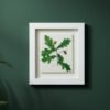 The Native Irish Fern Leaf – Original Ceramic Wall Art is showcased on a dark green wall in a white frame, with lush green plants partially visible at the edges of the image.