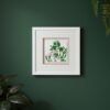 The "Large Colourful Meadow Mix – Fine Art Print" hangs on a dark green wall, featuring pink and white cows with green leaves, surrounded by lush, leafy plants.