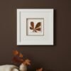 A fine art print, "Large Colourful Meadow Mix," hangs on a dark brown wall above a wooden table adorned with a vase of autumn leaves and a cream knitted blanket.