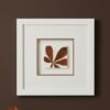 The "Large Colourful Meadow Mix – Fine Art Print" is displayed in a white square frame on a dark brown wall, featuring pressed brown leaves arranged in a fan shape against a white background, with autumn leaves visible in the lower left corner.