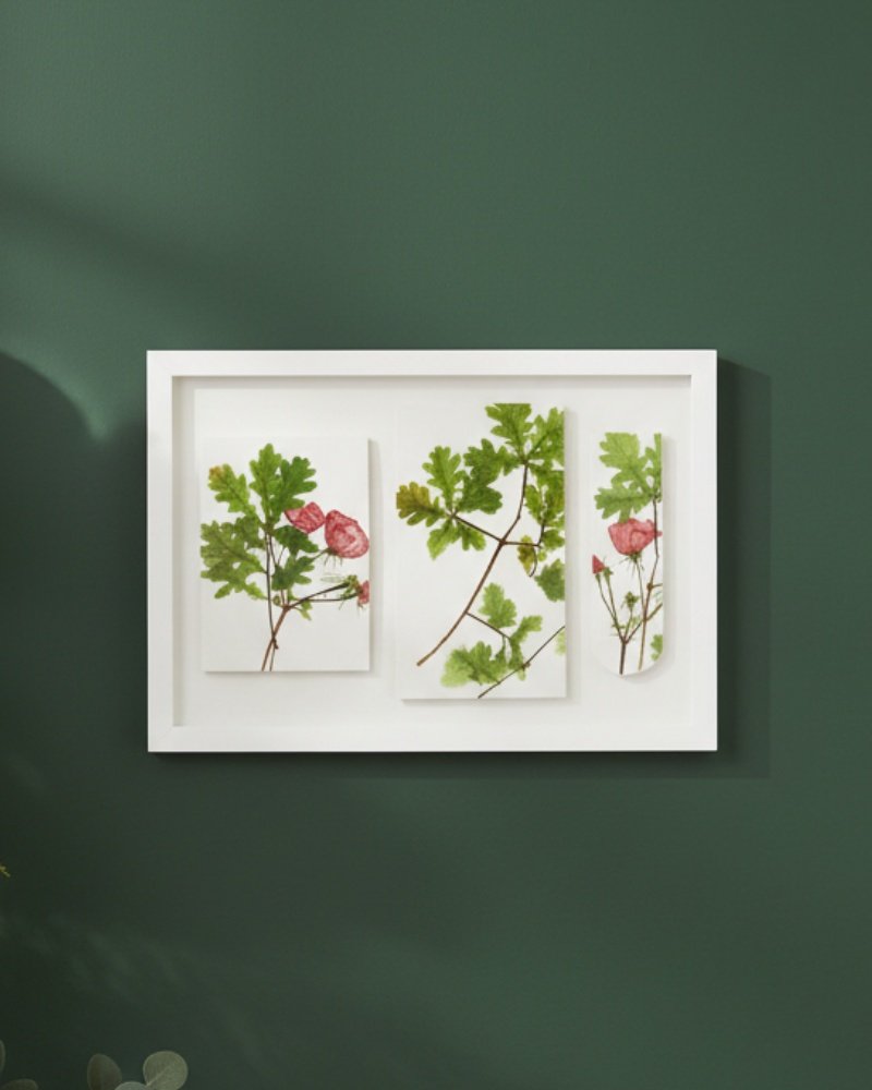 The Large Colourful Meadow Mix – Fine Art Print features three panels with botanical illustrations of green leaves and pink flowers. Displayed in a white frame on a dark green wall, it offers a modern, minimalist touch to your space.