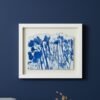 Wild Meadow Mix (Medium), a framed artwork by an Irish artist with blue botanical silhouettes on white paper, is displayed on a dark blue wall above a gray couch and a small vase of dried grass.