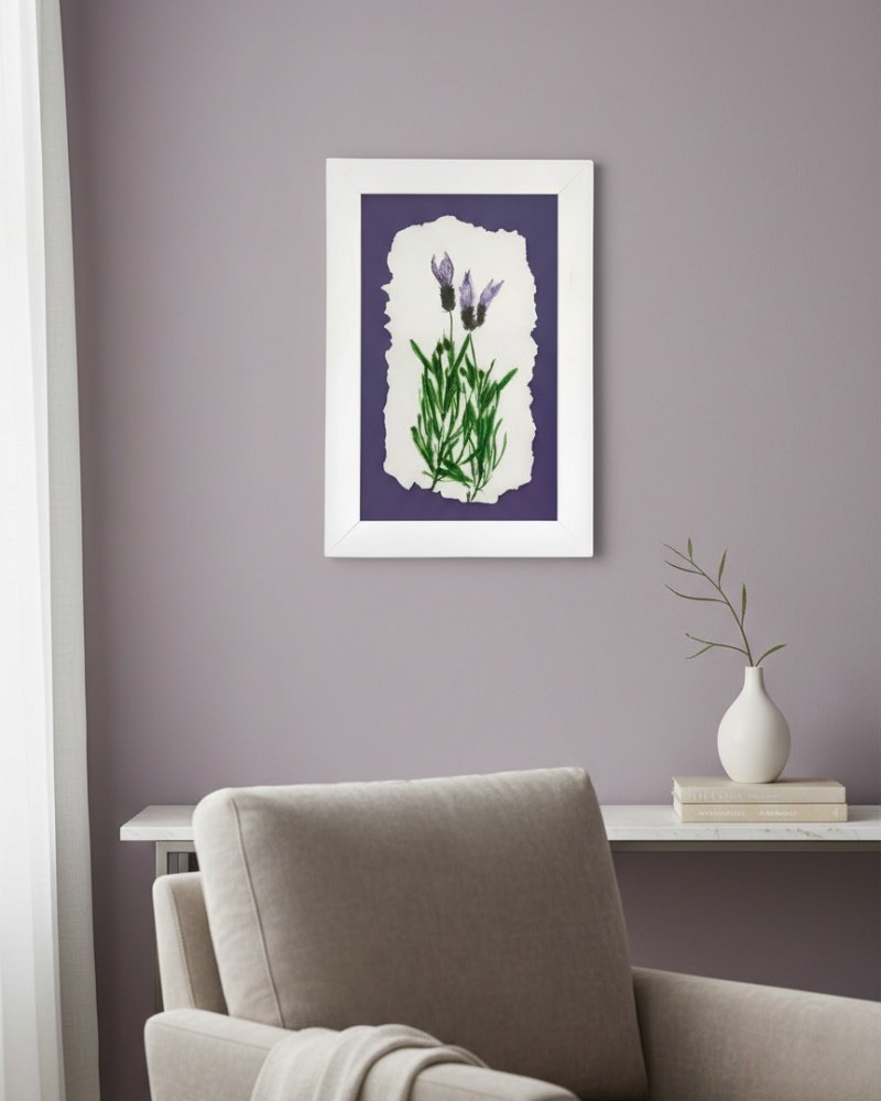 A minimalist room with a light gray armchair, white desk holding a vase with a branch, and "Wild Meadow Mix (Medium)"—a framed botanical artwork of purple flowers by an Irish artist—on a lavender wall, bathed in natural light from the left.