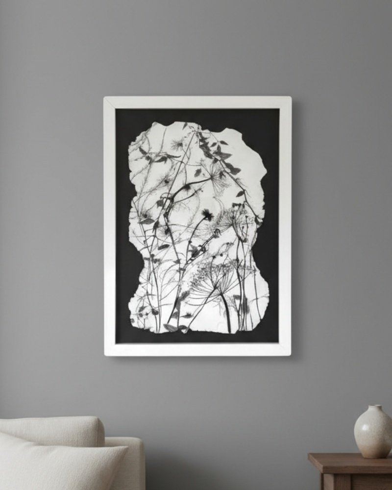 The Wild Meadow Mix (Medium), a framed black-and-white botanical artwork by an Irish artist, hangs above a wooden table and beside a light sofa on a gray wall, displaying silhouettes of delicate meadow plants and flowers.