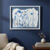 The "Rosemary – Original Ceramic Wall Art," featuring blue wildflowers on white, is displayed on a dark blue wall above a beige armchair with a blanket, beside a wooden side table holding books and a dried floral arrangement.