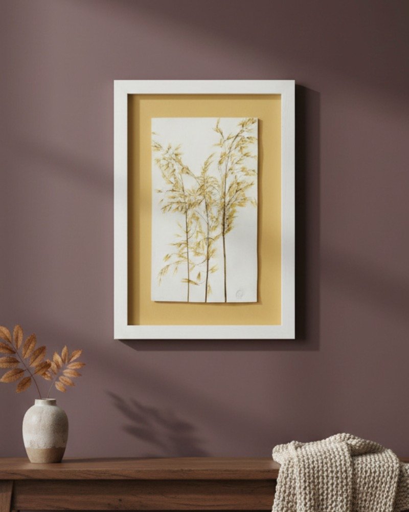 The Rosemary – Original Ceramic Wall Art, inspired by botanical Irish themes, features pressed grasses on yellow ceramic and hangs on a mauve wall above a wooden surface with a small vase of dried sprigs and a textured beige blanket.
