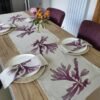 A wooden dining table set with the Purple Dulse Linen Table Runner, matching placemats and napkins, white plates, and a vase of colorful flowers in the center. Four purple chairs surround the table.
