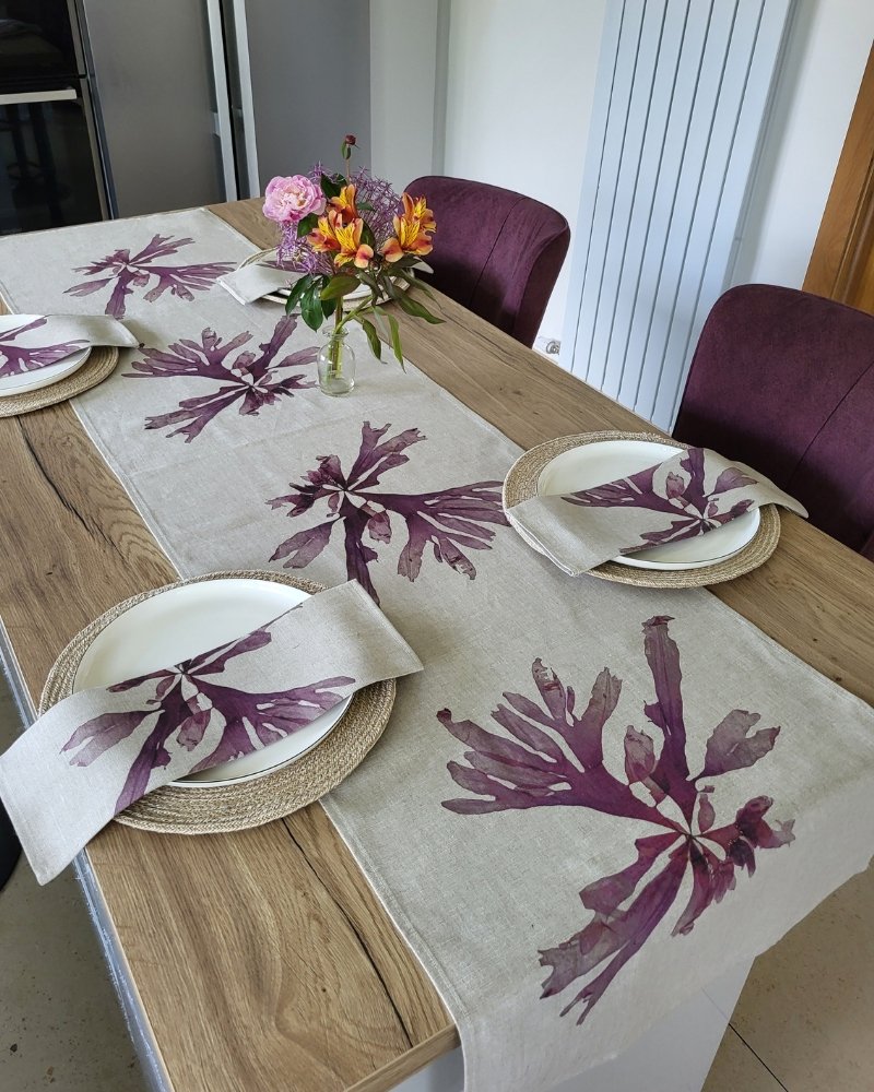 A wooden dining table set with the Purple Dulse Linen Table Runner, matching placemats and napkins, white plates, and a vase of colorful flowers in the center. Four purple chairs surround the table.