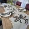 A wooden dining table set with four place settings, each featuring a woven placemat, a plate, and a folded napkin. A floral centerpiece adds elegance, while the Brown Dulse Seaweed Linen Table Runner with its leaf designs beautifully decorates the table.