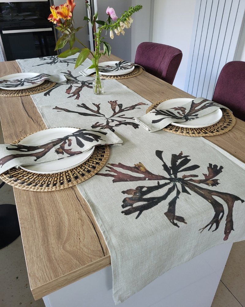 A wooden dining table set with four place settings, each featuring a woven placemat, a plate, and a folded napkin. A floral centerpiece adds elegance, while the Brown Dulse Seaweed Linen Table Runner with its leaf designs beautifully decorates the table.