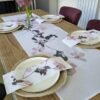 A wooden dining table set for four is adorned with floral-patterned placemats, napkins, and the Climbing Clematis Linen Table Runner. Each place setting features a white plate atop a woven placemat, and a vase of purple flowers serves as an elegant centerpiece.