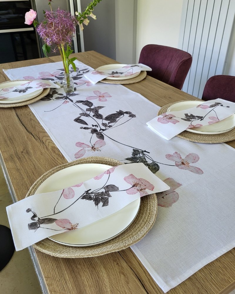 A wooden dining table set for four is adorned with floral-patterned placemats, napkins, and the Climbing Clematis Linen Table Runner. Each place setting features a white plate atop a woven placemat, and a vase of purple flowers serves as an elegant centerpiece.