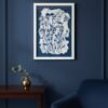 The Tall Wild Meadow Mix framed print, featuring blue and black flower outlines inspired by Irish art, hangs on a dark blue wall above a wooden table with a gold vase and green sprig. A navy blue chair is partially visible in front.