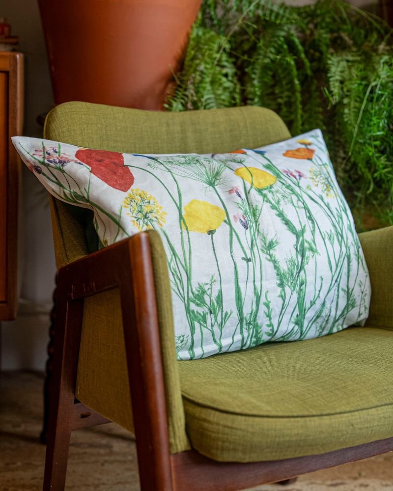 A green mid-century modern armchair holds our Colourful Poppy Mix Linen Cushion, displaying a vibrant wildflower print. The cozy room is accented with leafy plants and a large terra cotta pot in the background.