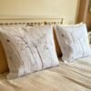 Two Coastal Seedheads Linen Cushions featuring delicate seedhead designs rest against a white headboard on a neatly made bed with beige bedding, evoking a serene coastal ambiance in the softly lit bedroom.