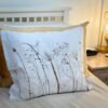 The Coastal Seedheads Linen Cushion, in white linen with a brown botanical seedhead print, is displayed on a bed with beige and white bedding. A nightstand holding a lamp and clock sits beside the bed.