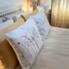 Two decorative pillows, including the Coastal Seedheads Linen Cushion embellished with a delicate botanical print, rest on a neatly made bed dressed in cream and beige bedding. Sunlight filters through patterned curtains, illuminating a lamp that sits on the bedside table in the background.