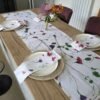 A wooden dining table set for four features white floral napkins, a Wild Sweet Pea Linen Table Runner, round placemats, white plates, and two vases of colorful flowers as the centerpiece.