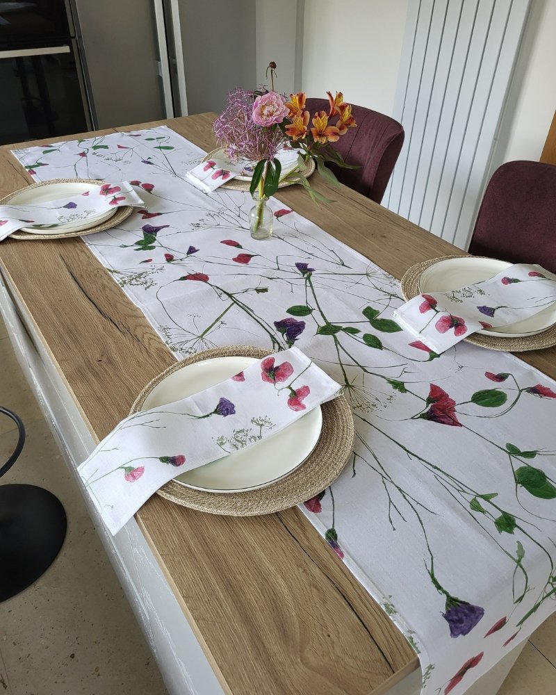 A wooden dining table set for four features white floral napkins, a Wild Sweet Pea Linen Table Runner, round placemats, white plates, and two vases of colorful flowers as the centerpiece.
