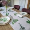 A wooden dining table set for four is adorned with woven placemats, white plates, and napkins — each decorated with green botanical prints. The centerpiece is the Garden Herbs Linen Table Runner, featuring leafy designs inspired by garden herbs, while a vase displays purple allium flowers.