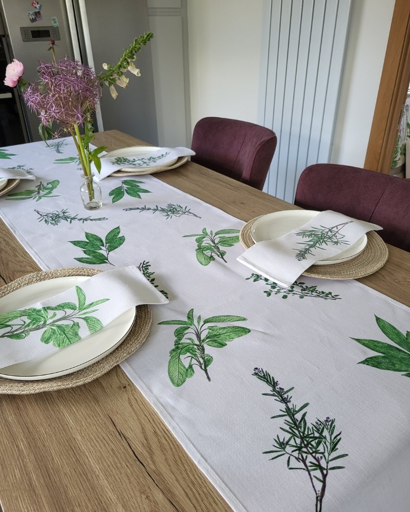 A wooden dining table set for four is adorned with woven placemats, white plates, and napkins — each decorated with green botanical prints. The centerpiece is the Garden Herbs Linen Table Runner, featuring leafy designs inspired by garden herbs, while a vase displays purple allium flowers.