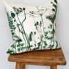 The Botanical Meadow Cushion in Green, featuring vibrant green prints of wildflowers and ferns on a crisp white background, is elegantly placed on a simple wooden stool against a plain white backdrop.