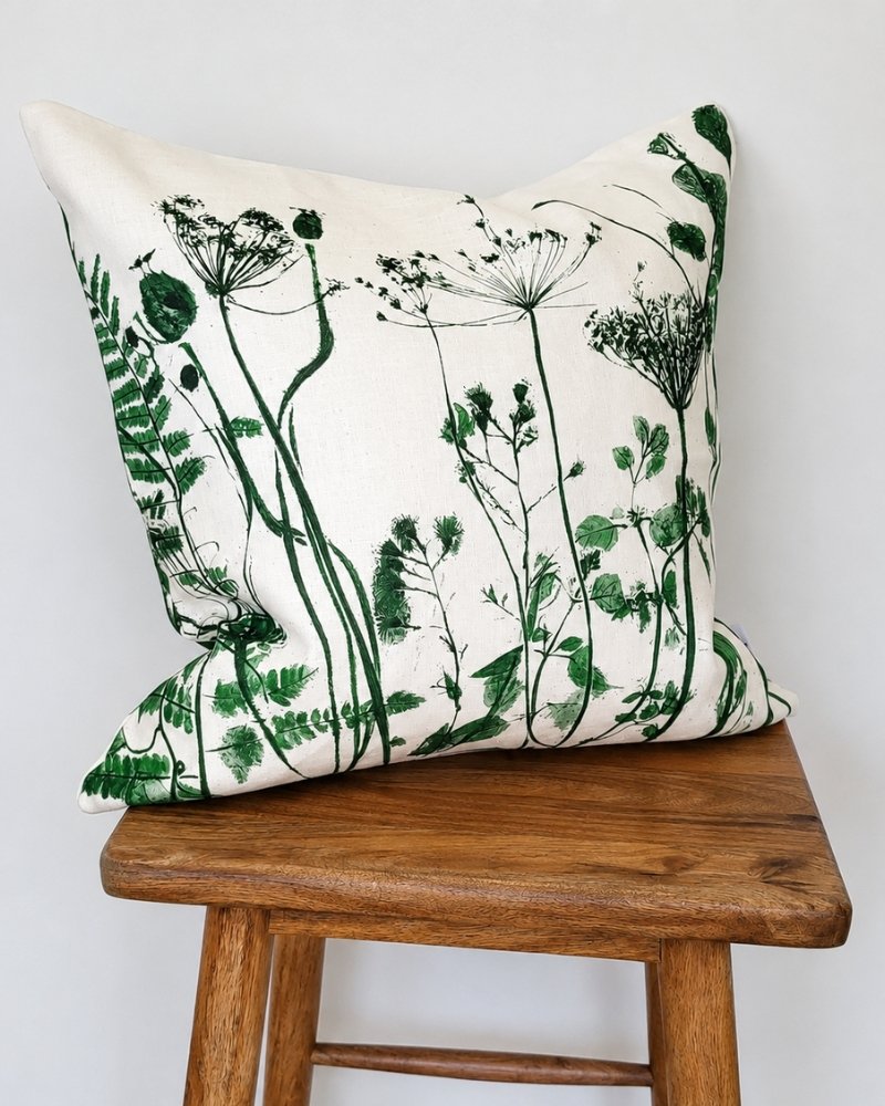 The Botanical Meadow Cushion in Green, featuring vibrant green prints of wildflowers and ferns on a crisp white background, is elegantly placed on a simple wooden stool against a plain white backdrop.