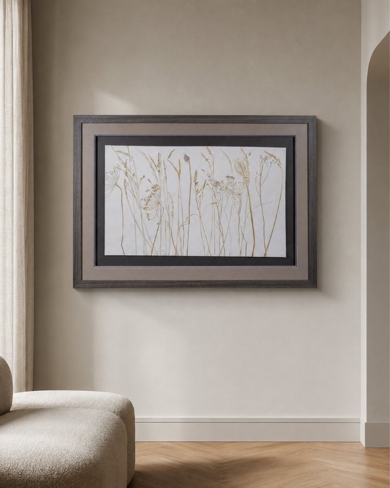 A framed print of “Coastal Seedheads” hangs on a light beige wall above a curved, light-colored chair in a minimalist room with wood flooring.