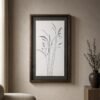 The “Grasses in Charcoal” framed minimalist botanical artwork hangs on a beige wall near a window, accented by a round chair and a wooden table topped with a dark vase filled with greenery.