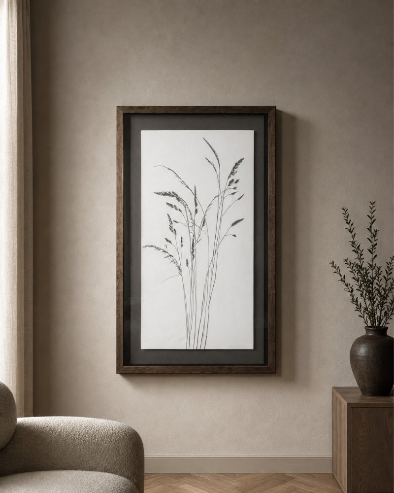 The “Grasses in Charcoal” framed minimalist botanical artwork hangs on a beige wall near a window, accented by a round chair and a wooden table topped with a dark vase filled with greenery.