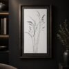 The framed Grasses in Charcoal minimalist line drawing hangs on a dark wall. Nearby, shelves display books and pottery, while a vase filled with green foliage sits on a table, creating a modern and elegant atmosphere.